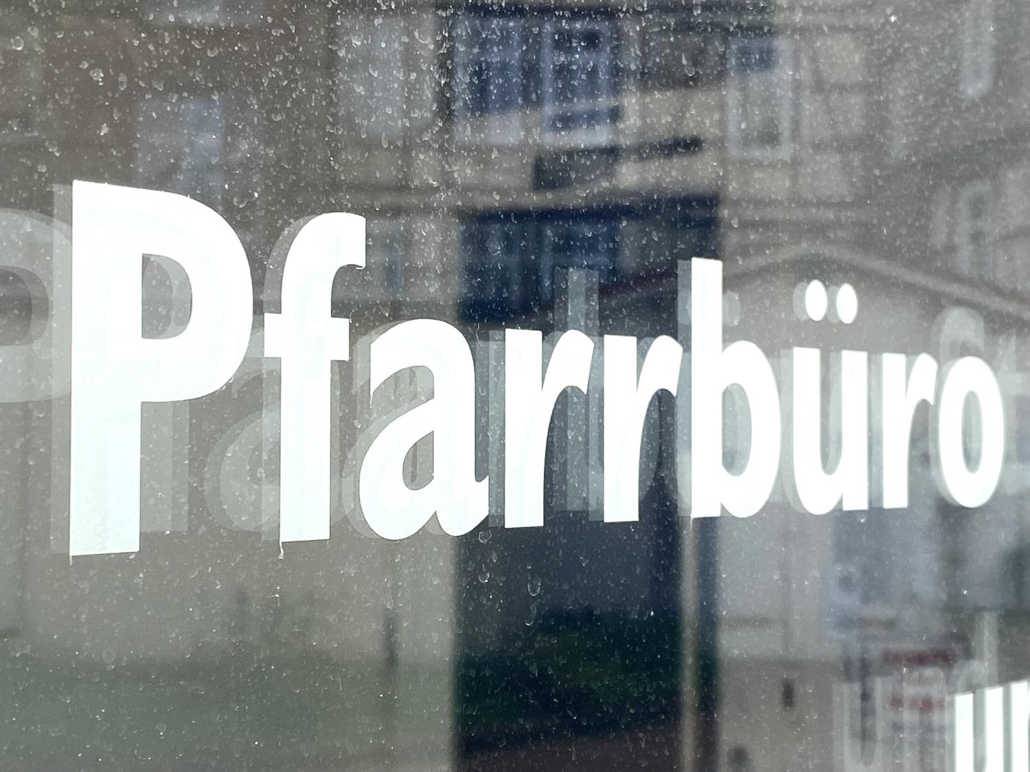 Weiße Schrift „Pfarrbüro“ auf einer Glasscheibe mit Spiegelungen. 